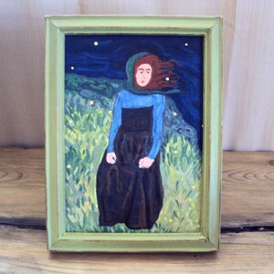 Op de afbeelding: Een ingelijst schilderij van een vrouw met rood haar die een blauwe en groene jurk draagt en in een veld met groen gras en gele bloemen staat. De achtergrond is een donkerblauwe lucht met witte sterren.