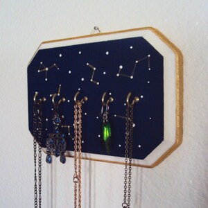 May include: A dark blue wooden jewelry organizer with gold trim and white painted stars. The organizer has five hooks for hanging necklaces and earrings. The hooks are adorned with gold-colored chains and a green beetle charm.