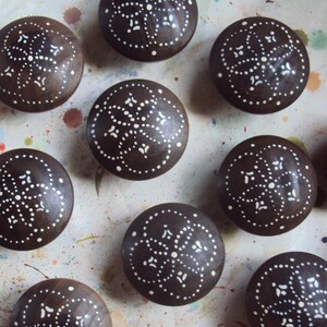 May include: Nine round wooden knobs with a dark brown background and white polka dots and swirls. The knobs have a decorative design.