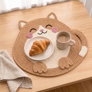 May include: A tan and white crocheted cat-shaped placemat featuring pink cheeks, pointed ears, and a curved tail. A small white plate holding a golden-brown croissant and a beige ceramic mug filled with coffee sit on the placemat surface.