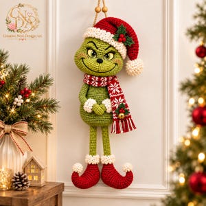 May include: A crocheted green character wall hanging featuring a red and white Santa hat, a matching red and white patterned scarf with a small gold bell, and red curled elf-style shoes with white pom-poms. The figure has a smiling expression.