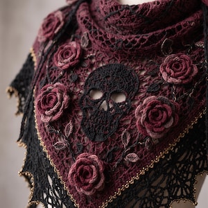 May include: A triangular crochet shawl featuring a central black skull motif surrounded by three-dimensional dark red roses and vine patterns. The shawl is crafted from deep burgundy and black yarn with a delicate openwork lace border and gold beads.