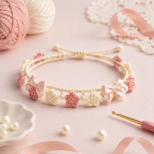 May include: A delicate handmade bracelet featuring a cream-colored crocheted cord adorned with small pink and cream crocheted flowers, white faux pearls, and two light pink satin ribbon bows, finished with a sliding knot closure mechanism.
