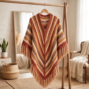 May include: A V-neck crochet poncho featuring a chevron pattern with alternating stripes in shades of rust orange, brown, cream, and tan. The garment is finished with long, dangling fringe along the bottom hem and is displayed on a wooden hanger.