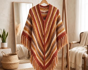 May include: A V-neck crochet poncho featuring a chevron pattern with alternating stripes in shades of rust orange, brown, cream, and tan. The garment is finished with long, dangling fringe along the bottom hem and is displayed on a wooden hanger.