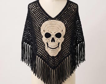 May include: A black crochet poncho featuring a large, cream-colored skull motif centered on the front. The garment has a V-neckline, an open-weave mesh construction, and long, dangling fringe trim along the bottom hemline and edges.