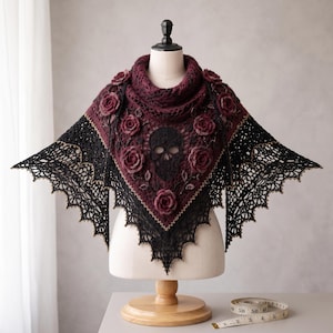 May include: A triangular crochet shawl featuring a deep burgundy center with a black skull motif and dark red roses, transitioning into a black lace border with gold-toned edges. A white measuring tape shows a length of 60 inches on the table.