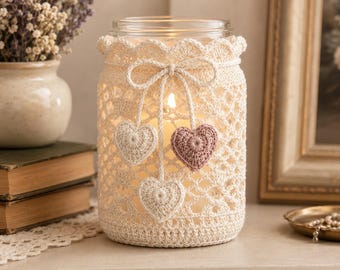 Puede incluir: Un frasco de vidrio transparente con una funda de ganchillo color crema que presenta un borde superior festoneado y un lazo decorativo. Tres pequeños corazones de ganchillo, dos en crema y uno en rosa empolvado, cuelgan del lazo.