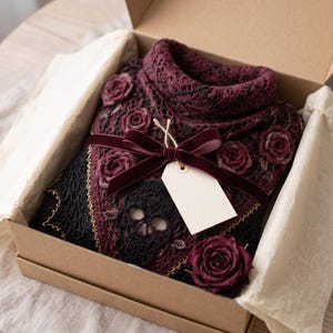May include: A dark burgundy and black crocheted sweater featuring intricate skull motifs and three-dimensional rose appliqu&eacute;s. The garment is neatly folded inside a cardboard gift box, tied with a deep red velvet ribbon and a blank white tag.