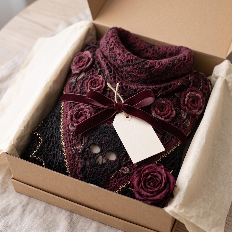 May include: A dark burgundy and black crocheted sweater featuring intricate skull motifs and three-dimensional rose appliqu&eacute;s. The garment is neatly folded inside a cardboard gift box, tied with a deep red velvet ribbon and a blank white tag.