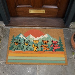 Puede incluir: Un felpudo rectangular de fibra de coco con un estampado gráfico de cinco osos bailando en colores verde, amarillo, naranja y rojo. El diseño incluye una cordillera, un gran sol naranja y árboles de hoja perenne, con rayas horizontales abajo.