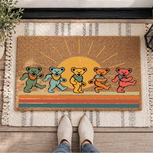 Pode incluir: Um capacho retangular de fibra de coco com um gráfico de cinco ursos dançantes em verde, azul claro, amarelo, laranja e rosa. Atrás dos ursos há um sol amarelo com raios, e abaixo deles três faixas horizontais em laranja, vermelho e azul.