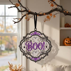 May include: A decorative hanging ornament featuring a black scalloped frame with a white honeycomb mesh background. The design includes two white ghosts, black spiderwebs, and the purple text BOO! with purple floral accents at the top and bottom.
