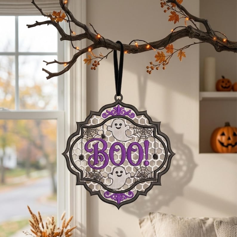 May include: A decorative hanging ornament featuring a black scalloped frame with a white honeycomb mesh background. The design includes two white ghosts, black spiderwebs, and the purple text BOO! with purple floral accents at the top and bottom.
