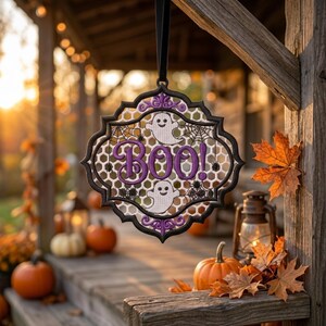 May include: A decorative hanging ornament with a black frame and a white honeycomb mesh center. It features purple embroidered text that reads BOO!, two white ghost figures, and black spiderweb designs. The ornament hangs from a black ribbon.