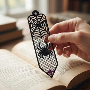 May include: A black embroidered bookmark featuring a spider web pattern at the top, a central spider motif, and a small purple fleur-de-lis accent at the pointed bottom. The bookmark has a circular hole at the top and a grid-like openwork design.