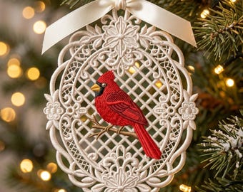 May include: An oval-shaped white lace ornament featuring an embroidered red cardinal bird perched on a brown branch. The intricate lace frame has a lattice pattern and floral details, topped with a white satin ribbon bow for hanging purposes.