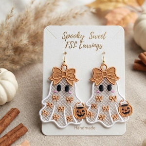 May include: A pair of white ghost-shaped earrings with gold-colored bow embroidery, black eyes, pink cheeks, and a small orange jack-o'-lantern bucket. The display card reads Spooky Sweet FSL Earrings and Handmade in gold script lettering.