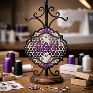 May include: A black metal stand holds a decorative hanging ornament featuring a white honeycomb pattern, two white ghosts, and the purple embroidered text "BOO!". The ornament is framed by a black scalloped border with purple accents and spiderwebs.