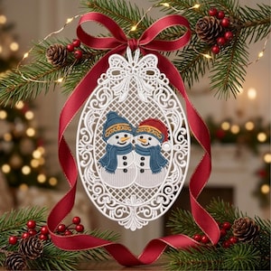 May include: An oval white lace ornament featuring two embroidered snowmen wearing blue scarves and hats, one with a blue hat and one with a red hat. The ornament is accented with a decorative white lace border and a deep red satin ribbon bow.