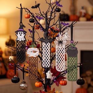 May include: Five vertical hanging ornaments with lattice patterns, featuring a ghost with a purple hat, a mummy pumpkin, a black bat, a white spider web with a spider, and a green cauldron with bubbling liquid, all suspended by black ribbons.