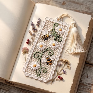 以下が含まれることがあります： 白いレースの長方形のしおり。ヒナギク、緑の茎、2匹のマルハナバチの刺繍が施されています。縁はスカラップ状で、上部にはクリーム色のタッセルが付いており、開いた本の上に置かれています。