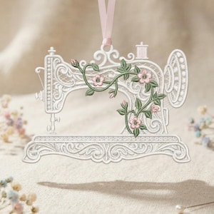 Può includere: Un ornamento bianco da appendere, simile al pizzo, a forma di macchina da cucire vintage. Il design presenta intricati motivi a volute ed è decorato con viticci verdi ricamati e piccoli fiori rosa. Un nastro rosa chiaro è fissato in alto.