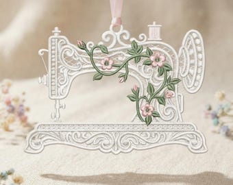 May include: A white, lace-like hanging ornament shaped like a vintage sewing machine. The design features intricate scrollwork and is decorated with embroidered green vines and small pink flowers. A light pink ribbon is attached to the top for hanging.