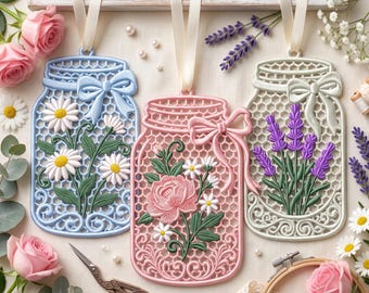 May include: Three hanging mason jar ornaments featuring intricate lattice patterns and floral designs. The left jar is light blue with daisies, the center is pink with a rose and daisies, and the right is cream with lavender. Each has a satin ribbon.