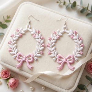 Puede incluir: Un par de pendientes colgantes con un diseño de corona circular hecho de hojas bordadas en blanco con pequeños detalles en rosa. En la base de cada corona hay un lazo bordado en rosa claro. Los pendientes cuelgan de ganchos de metal plateado.