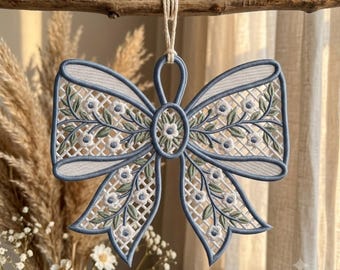 May include: A decorative bow ornament featuring a blue embroidered border and a white lattice mesh center. The design includes small white flowers with blue centers and green leaves embroidered across the surface, suspended by a white cord.