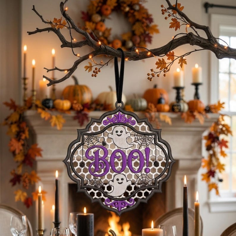 May include: A decorative hanging ornament with a black scalloped frame, featuring a white mesh background, two white ghost illustrations, black spiderweb patterns, and the word "BOO!" embroidered in purple thread, suspended by a black ribbon.