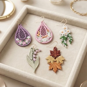 Puede incluir: Cinco pendientes colgantes con diseños detallados: una lágrima morada con flores, una lágrima rosa con detalles florales, un racimo de margaritas blancas con hojas verdes, una rama de lirio de los valles verde claro y tres hojas de arce superpuestas.