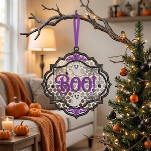 May include: A decorative hanging ornament featuring a black scalloped frame with a white honeycomb background, purple text reading BOO!, two white ghost illustrations, and black spiderweb accents, suspended by a purple ribbon loop.
