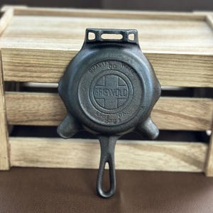 Puede incluir: Una sartén de hierro fundido negro Griswold con un mango largo y un lazo para colgar en la parte superior. La parte inferior presenta texto en relieve que dice Quality Ware, Griswold, Erie PA y 570A, centrado en un diseño de cruz.