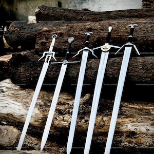 May include: Five metal swords with silver-toned blades and dark handles arranged in a row. Each sword features a distinct crossguard design, including one with a circular emblem and another with a unique pronged guard, all resting against logs.