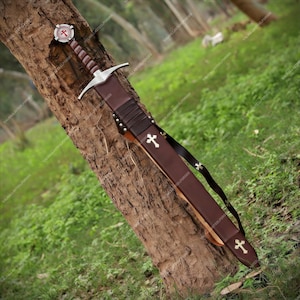 May include: A medieval-style sword with a brown leather-wrapped grip and a silver crossguard. The pommel features a circular emblem with a red cross. The brown leather scabbard is adorned with two silver cross emblems and includes a black leather belt.