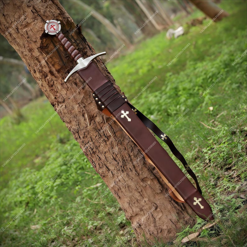 May include: A medieval-style sword with a brown leather-wrapped grip and a silver crossguard. The pommel features a circular emblem with a red cross. The brown leather scabbard is adorned with two silver cross emblems and includes a black leather belt.