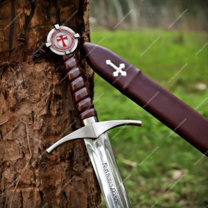 May include: A steel sword with a brown leather-wrapped grip and a circular pommel featuring a red cross emblem. The blade is engraved with the Latin text NON NOBIS DOMINE. A matching brown leather scabbard with a silver cross inlay is positioned nearby.