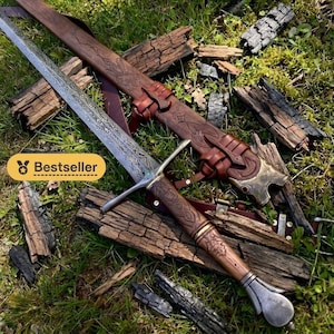 Könnte beinhalten: Ein Langschwert mit gemusterter Metallklinge, einem braunen Holzgriff mit kunstvollen Schnitzereien und einem silbernen Knauf. Das Schwert wird von einer passenden braunen Lederscheide mit geprägten Mustern und Metallakzenten begleitet.