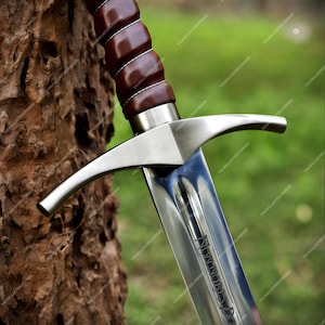 May include: A steel sword featuring a dark brown leather-wrapped grip with white stitching, a polished silver crossguard, and a blade with a central fuller. The blade is engraved with the text "NON NOBIS DOMINE" near the hilt area.