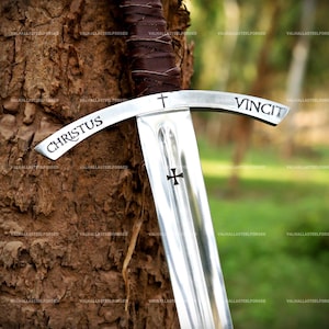 May include: A steel sword featuring a brown leather-wrapped hilt and a crossguard engraved with the text CHRISTUS VINCIT and a small cross symbol. The blade displays a central fuller and a black cross emblem etched into the polished metal surface.