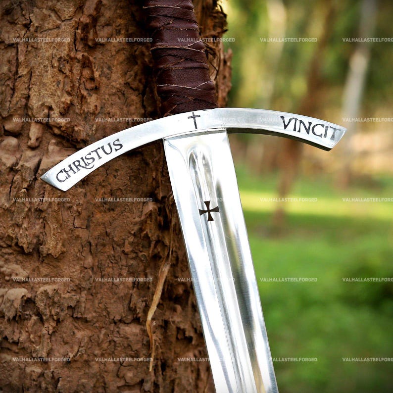 May include: A steel sword featuring a brown leather-wrapped hilt and a crossguard engraved with the text CHRISTUS VINCIT and a small cross symbol. The blade displays a central fuller and a black cross emblem etched into the polished metal surface.