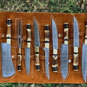 Puede incluir: Un juego de ocho utensilios de cocina que incluye un cuchillo de carnicero, un tenedor de trinchar y seis cuchillos con hojas de acero de Damasco y mangos de asta texturizados, organizados en una funda de cuero marrón enrollable con broches.