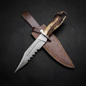 May include: A fixed-blade knife with a textured antler handle and a silver-toned metal blade featuring a serrated edge and engraved script. The knife is positioned over a brown leather sheath with visible stitching and a metal snap closure.