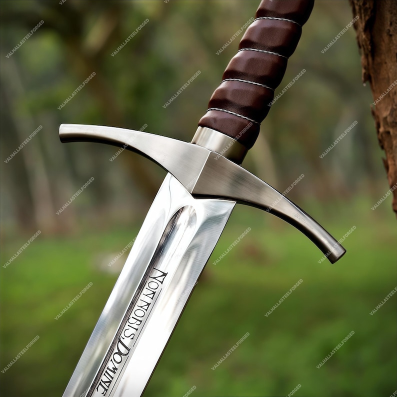 May include: A steel sword featuring a brown leather-wrapped grip with silver wire accents, a polished metal crossguard, and a blade engraved with the Latin text Non Nobis Domine. The blade has a central fuller running down its length.