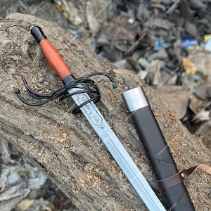 May include: A steel sword with a wooden handle and a complex black metal guard. The blade is engraved with the text 'Master of Fate' and is accompanied by a black scabbard featuring a silver-coloured metal tip and a brown leather strap.