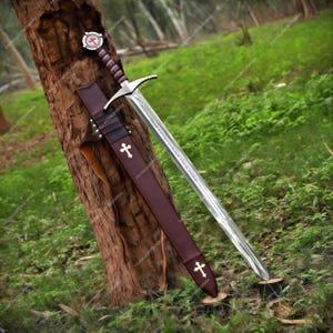 May include: A steel sword with a double-edged blade, a silver crossguard, and a dark brown leather-wrapped grip. The pommel features a circular emblem with a red cross. It includes a matching dark brown leather scabbard with two gold crosses.