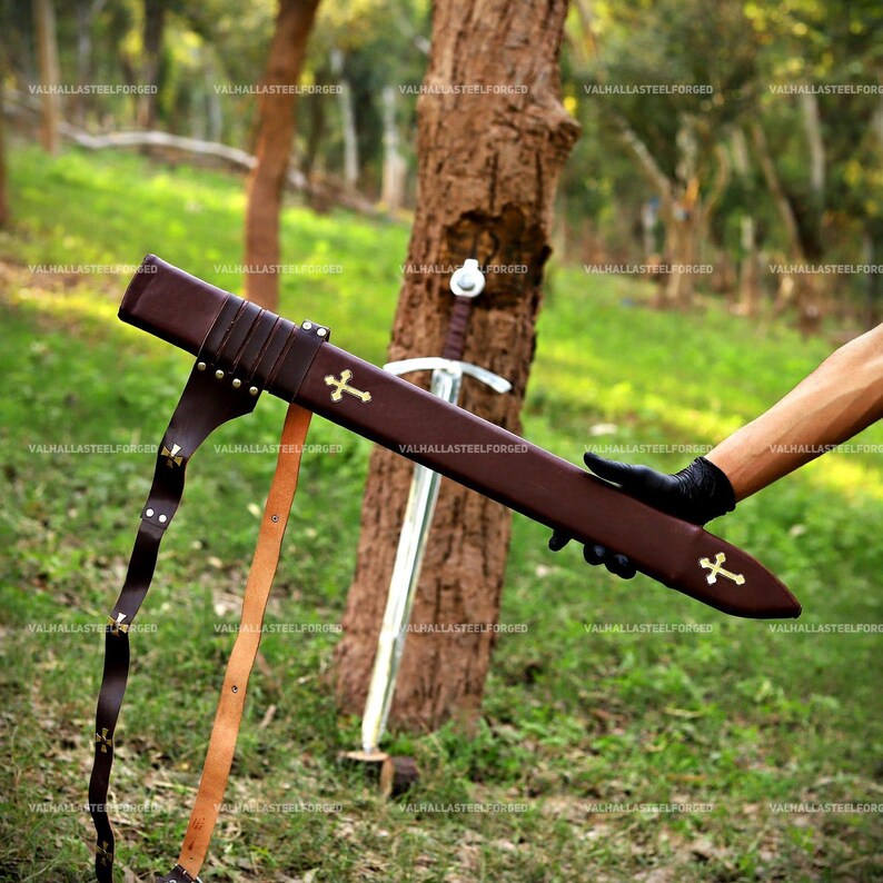 May include: A brown leather scabbard featuring two gold-colored cross emblems and a matching leather belt strap. The scabbard is held horizontally, while a metal sword with a cross-shaped hilt stands upright against a tree trunk in the background.