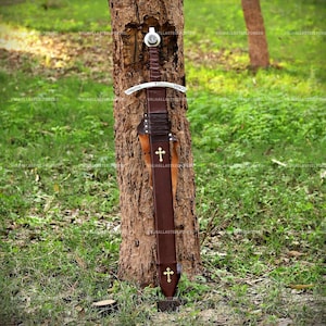 May include: A sword with a brown leather-wrapped hilt and a matching brown leather scabbard. The metal crossguard is engraved with the text CHRISTUS VINCIT. The scabbard features two gold-colored cross emblems, one near the top and one at the base.